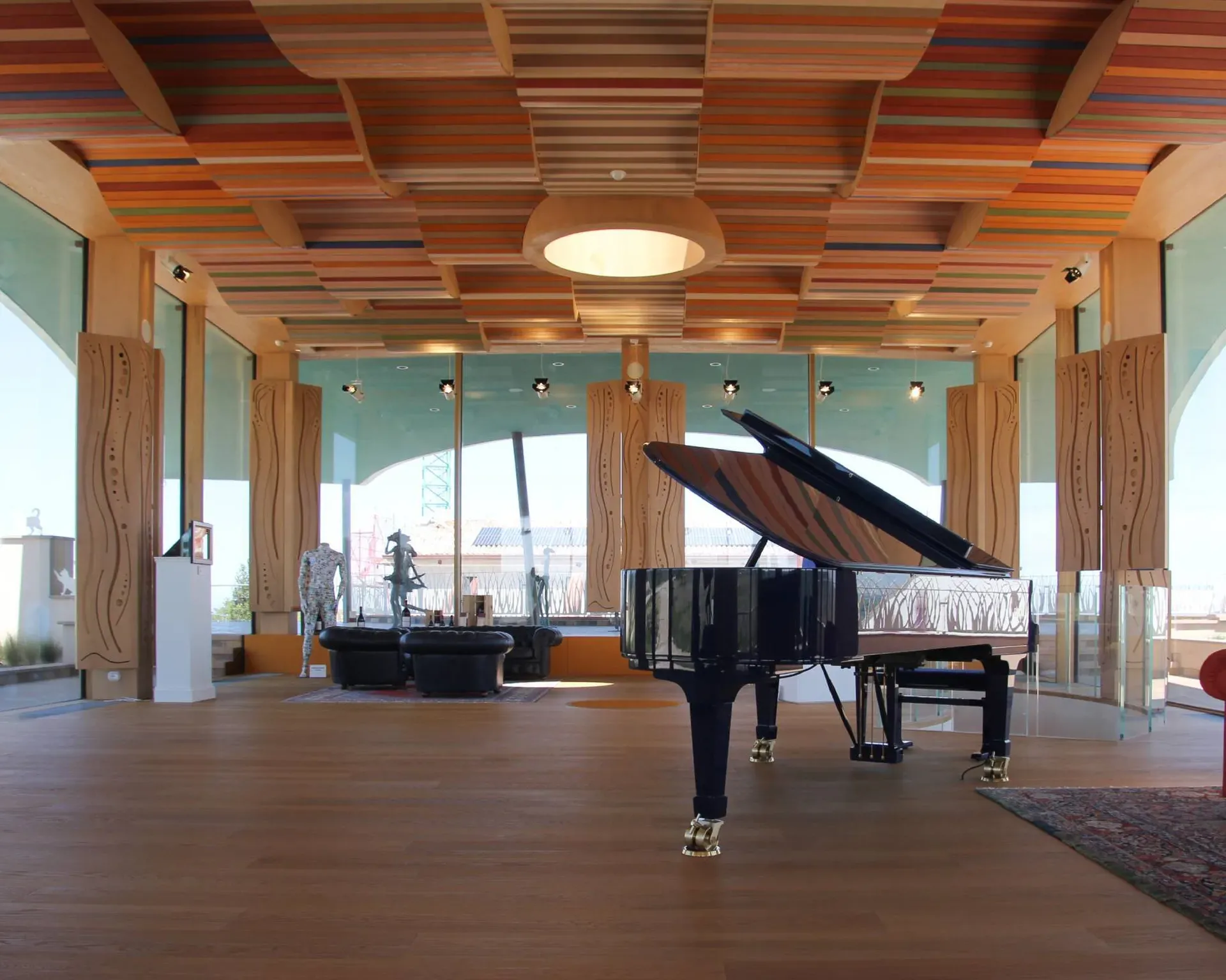Interno moderno con pianoforte a coda nero, coperchio aperto, soffitto a listelli colorati, colonne in legno e grandi vetrate; divano rosso in zona lounge.