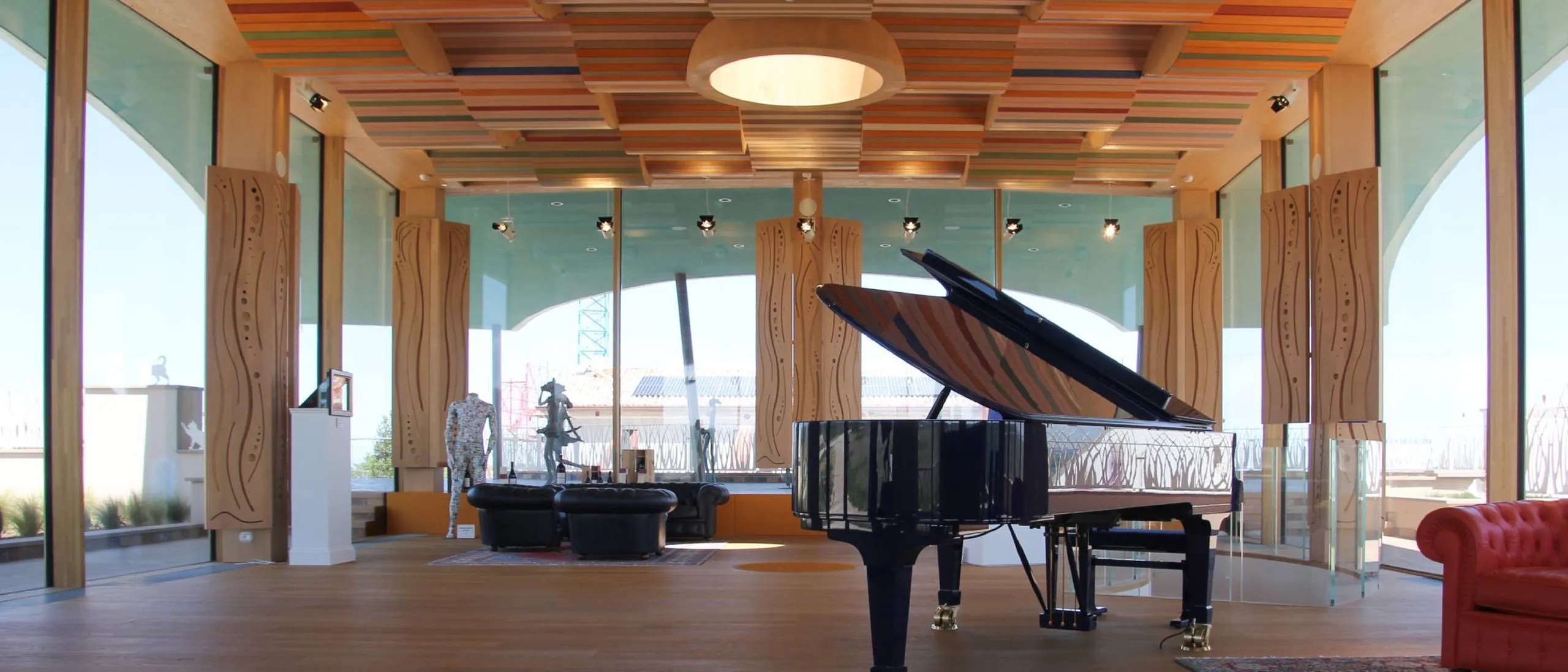 Interno moderno con pianoforte a coda nero, coperchio aperto, soffitto a listelli colorati, colonne in legno e grandi vetrate; divano rosso in zona lounge.