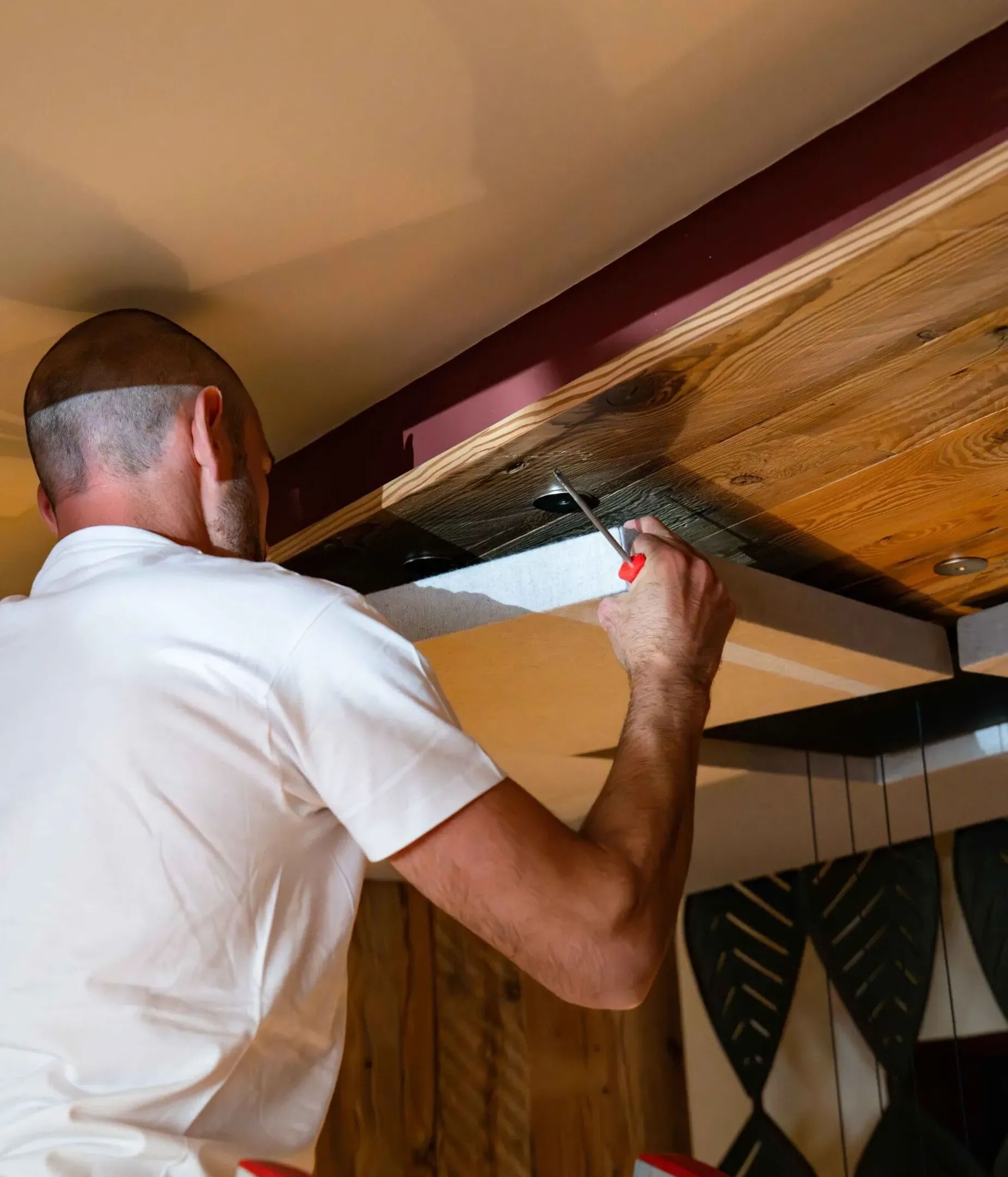 Una persona in camicia bianca su una scala lavora sul soffitto in legno, con un cacciavite per fissare un elemento; pareti con decorazioni scure.