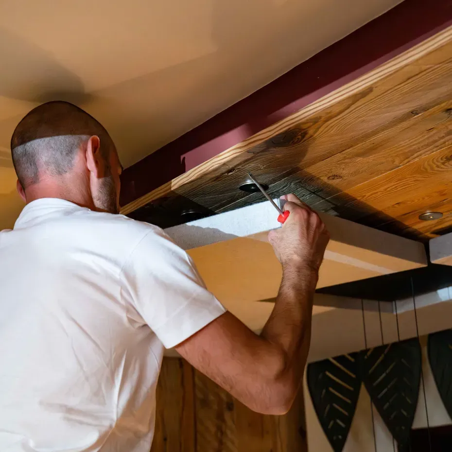 Una persona in camicia bianca su una scala lavora sul soffitto in legno, con un cacciavite per fissare un elemento; pareti con decorazioni scure.
