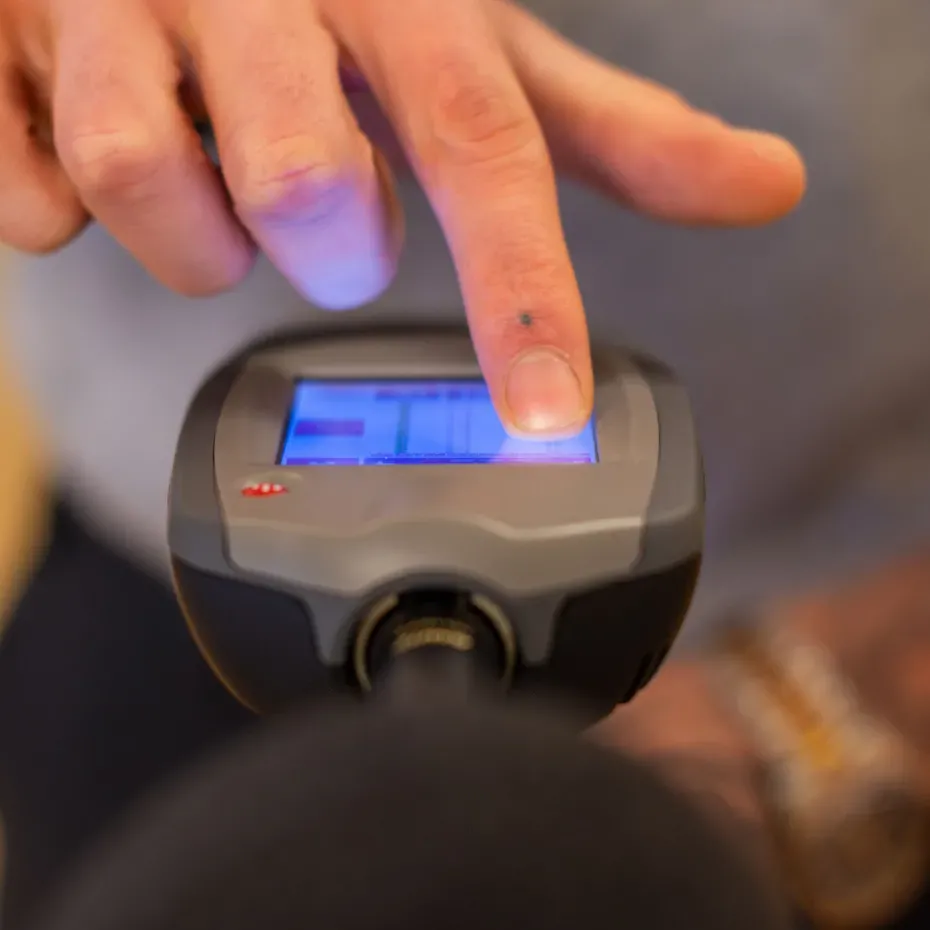 Mano che preme lo schermo blu di un misuratore portatile Silenzia, corpo grigio, durante una verifica acustica interna.