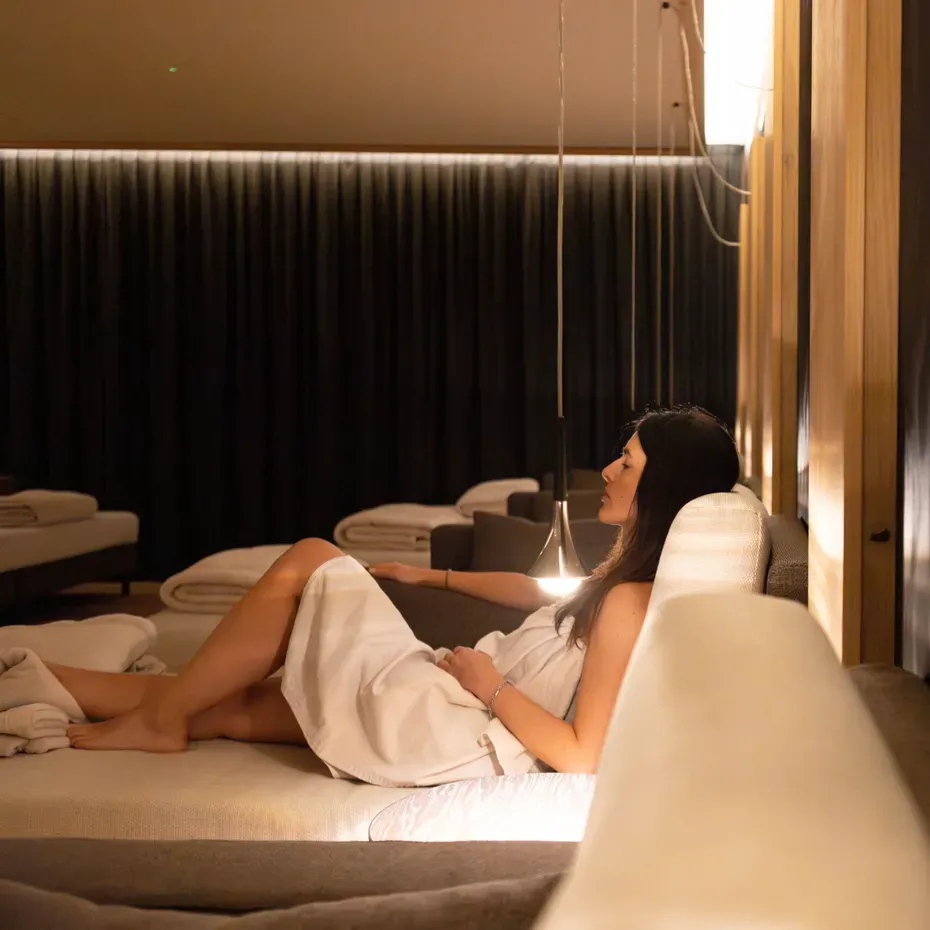 Persona in accappatoio bianco distesa su un divano in una lounge spa, luci soffuse e pannelli acustici creano un’atmosfera rilassante.