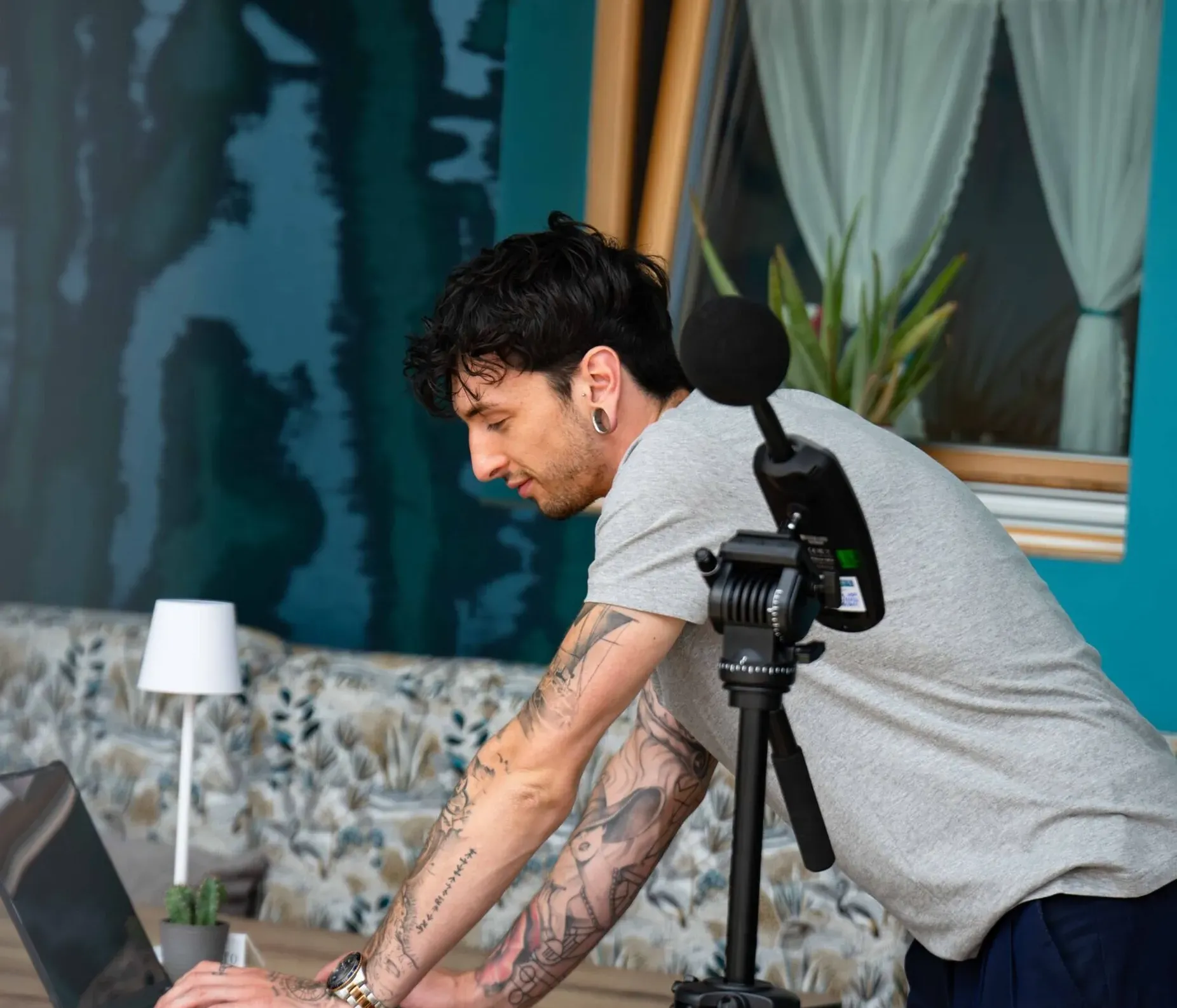 Persona con tatuaggi al tavolo lavora sul laptop, accanto a un microfono su treppiede; sfondo blu, finestra con tende, divano e pianta.