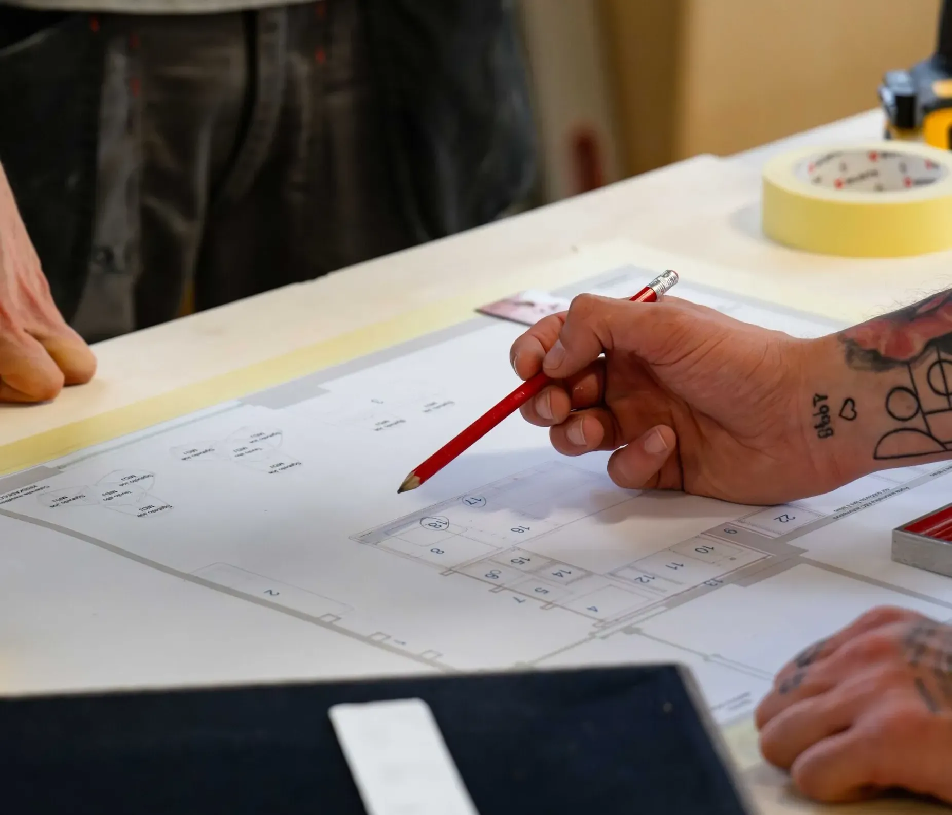 Due persone collaborano su una planimetria di progetto acustico: una mano tiene una matita rossa sopra schizzi, con attrezzi e nastro sul tavolo.