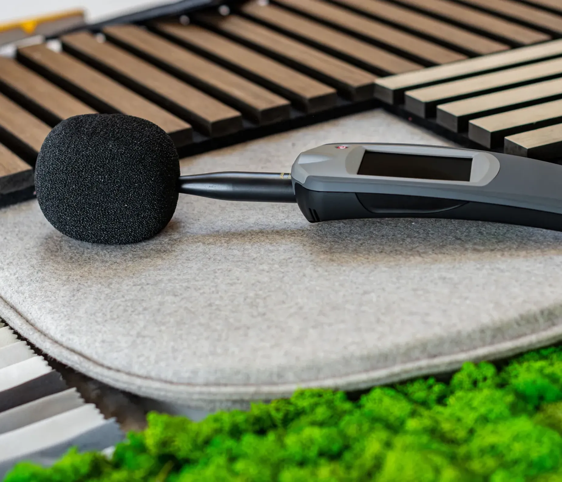 Microfono con copertura in schiuma su campioni acustici interni (tessuto, listelli di legno, moquette) con verde sullo sfondo.