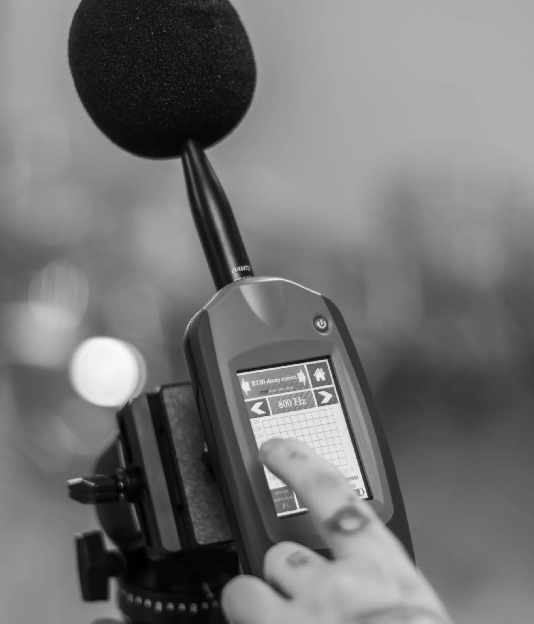 Black-and-white close-up of a microphone with foam windscreen on a stand and a handheld sound level meter, with a finger on the display during an acoustic measurement.