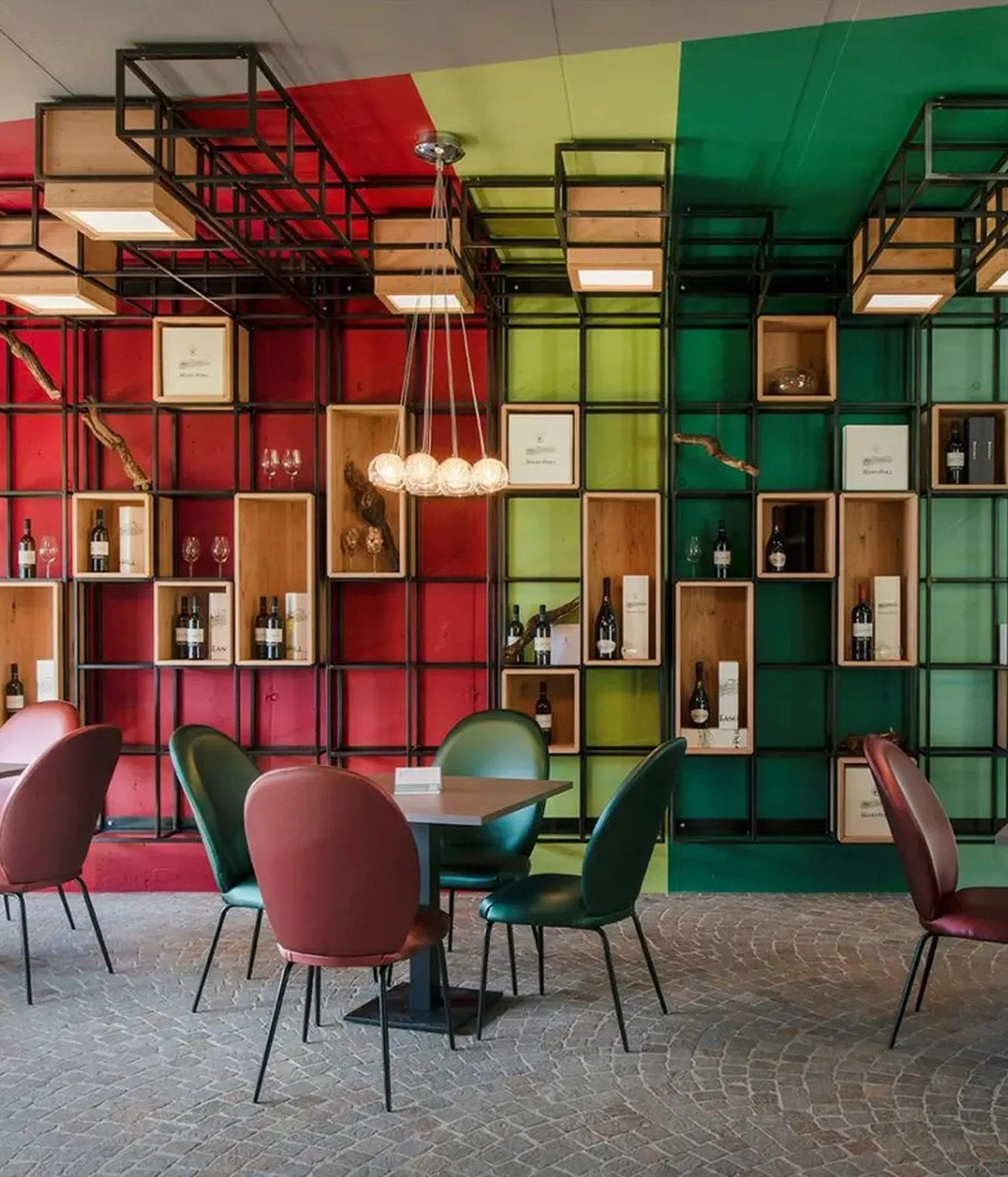 Interior with a multicolored grid-wall display of bottles and shelves, glass globe chandeliers, and round tables with red, green, and maroon chairs.