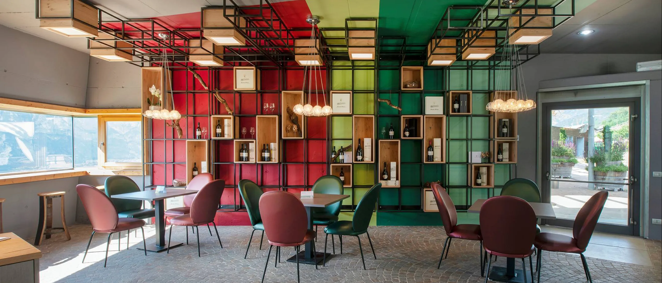 Interior with a multicolored grid-wall display of bottles and shelves, glass globe chandeliers, and round tables with red, green, and maroon chairs.