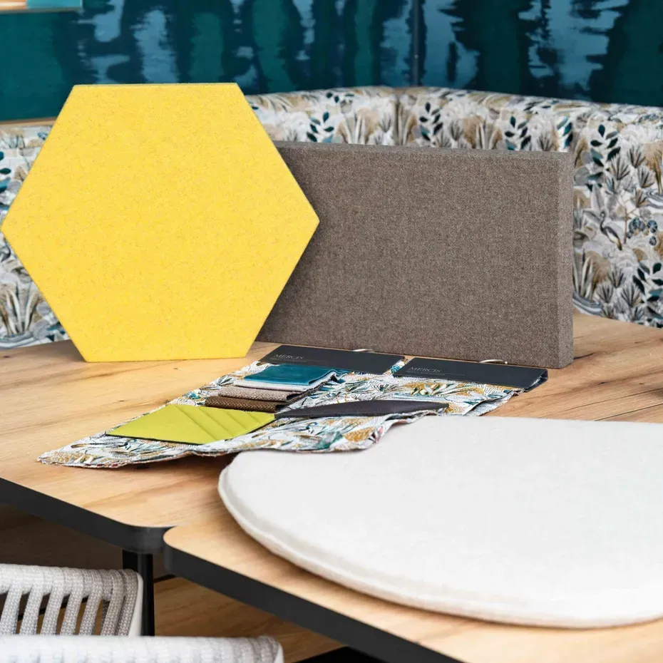 Set di pannelli acustici su tavolo in legno: esagono giallo, pannello grigio, campioni tessili; sfondo con divano floreale e parete verde-blu.